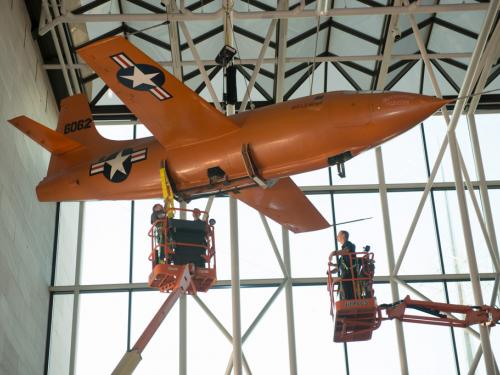 Two lifts are employed so that Museum employees can securely move the Bell X-1, an orange monoplane, from the ceiling to the floor of the Museum's Boeing Milestones of Flight Hall.