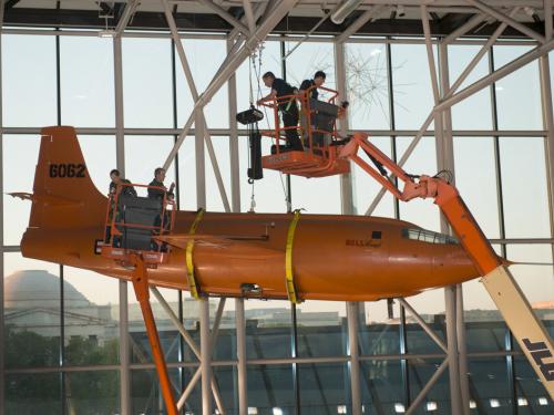 Using two lift machines, Museum employees work to secure the Bell X-1, an orange military monoplane, from the ceiling of the Boeing Milestones of Flight Hall onto the floor.