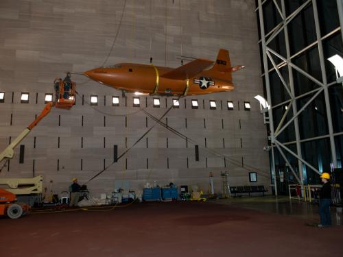 Museum employees in the middle of lowering the Bell X-1, an orange military monoplane, from the ceiling to the floor of the Boeing Milestones of Flight Hall. The aircraft is partially lowered.