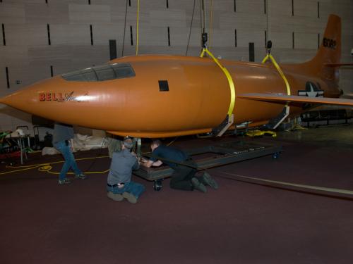 Museum employees secure the fully lowered Bell X-1, an orange military monoplane, as it stands stationary on the floor of the Boeing Milestones of Flight Hall.