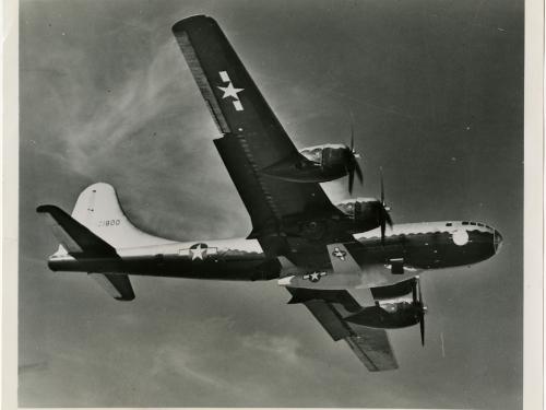 Bottom view of the Bell X-1, a military aircraft, being lifted under a B-29, a larger military aircraft. The B-29 is in flight.