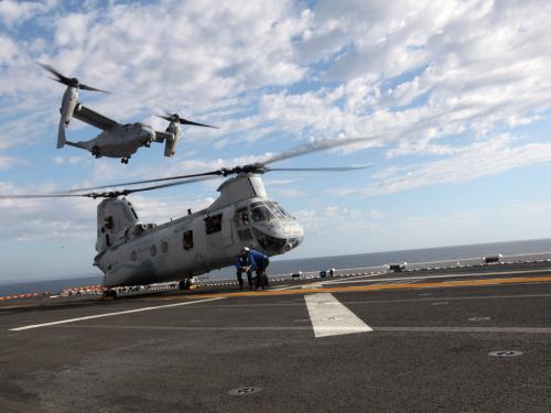 A helicopter with two sets of propellors placed on the sides of the aircraft flies over a helicopter on a tarmac with propellors on top of the body.