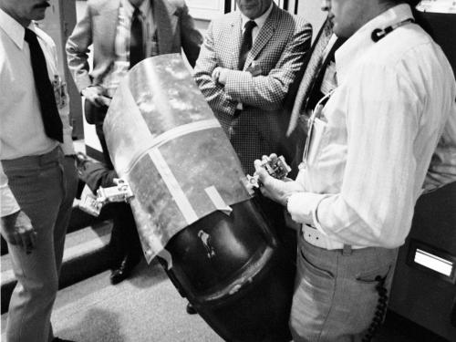 A group of five male NASA employees on Earth work together to come up with a solution for a wheel fender repair on the Lunar Module during the Apollo 17 mission.