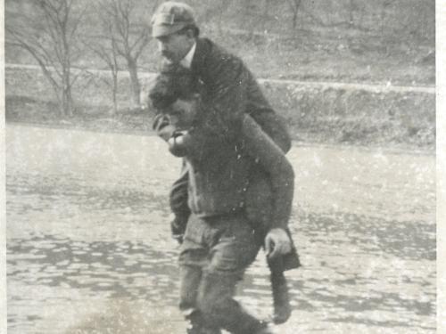 A mechanic working with Orville Wright gives Orville a piggyback as they walk in a river.