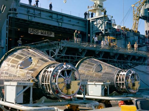 Frontal view of two conical-shaped spacecraft placed on their side as they are transported from a U.S. Navy carrier.