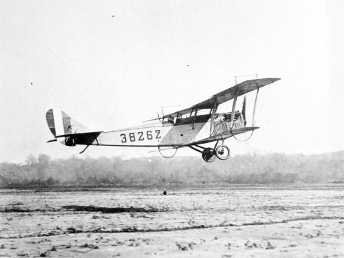 Side view of light-colored biplane in flight. Registration number "38262" written on back half of fuselage.