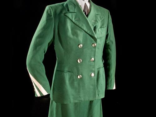 Front view of green stewardess uniform with green jacket with three pockets and six brass buttons, a green stewardess cap, and green skirt. White scarf worn on neck. Styled on mannequin.