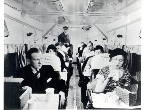 Interior of an early commercial aircraft. People sit in seats that have two rows of one seat apiece. The people in the front seem to have received some form of food service.