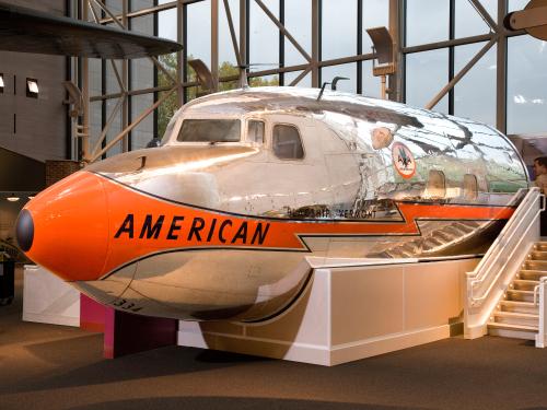 Nose, cockpit, and front fuselage section of a metal commercial airliner with orange, black, and silver-colored American Airlines livery.