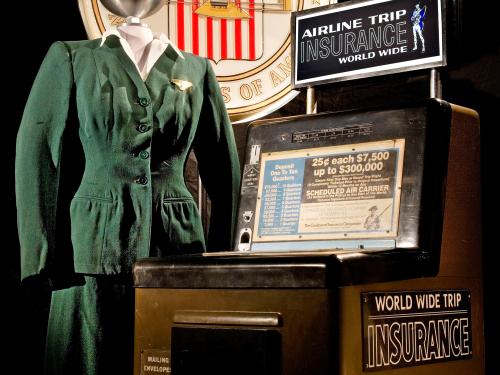 Brown-colored vending machine selling airline flight insurance. Next to the vending machine is a green flight attendant uniform styled on mannequin.