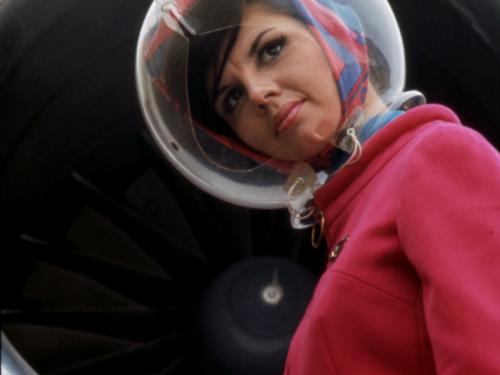 Side view of a flight attendant wearing a red flight uniform and a plastic, bubble-shaped helmet.