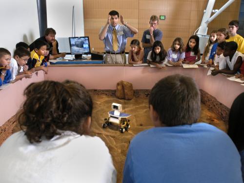 Museum visitors control a Mars rover model on a surface similar to the surface of Mars.