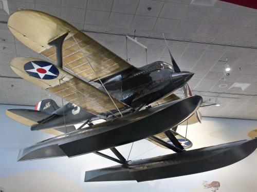 Black seaplane with pontoon floats instead of wheels and cream colored wings, hanging in museum