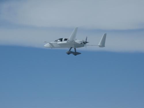 Side view of a white aircraft in flight with fixed landing gear and a propellor located on the rear of the fuselage.