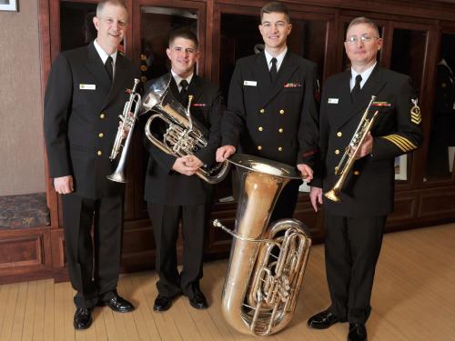 A set of four members of the U.S. Navy stand together in uniform, holding their respective brass instruments.