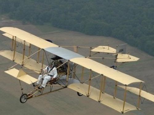 Diagonal side view of a biplane with yellow wings and a wooden frame during flight.