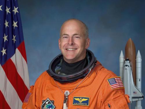 A portrait of a man in front of the american flag and a model of the space shuttle. He wears an orange astronaut suit and has an astronaut helmet in his hand.