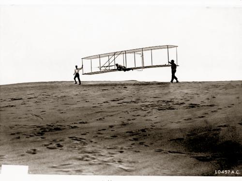 Orville Wright is prepared for takeoff in a test using the 1902 Wright Glider. The Wright Glider is being carried by Wilbur Wright and Dan Tate.