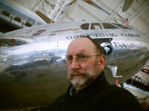 A white male archives specialist at the museum poses below a silver commercial airplane.