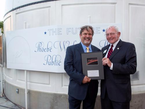 Two white men pose together with a dedication plaque as they stand outside of a white building.