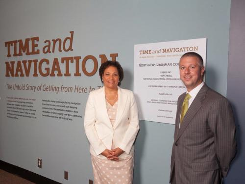 Two people, a woman and a male, stand next to the sponsorship plague for an exhibit that their corporation sponsored.