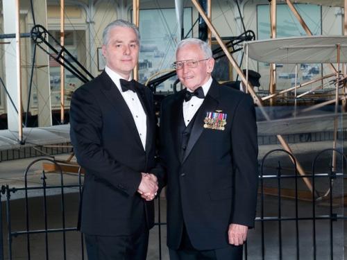 Two men stand together at an awards ceremony, with the left man shaking the hand of the right man.