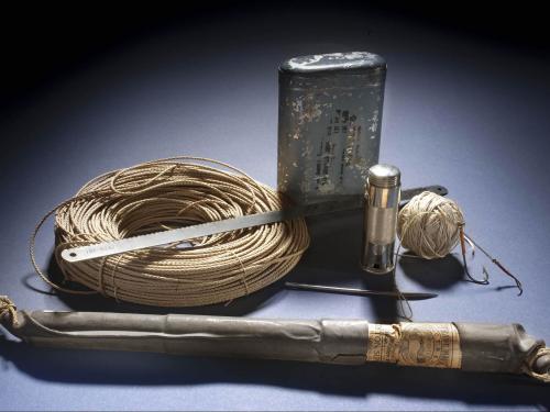 A set of emergency survival suplies including a fishing line and hook, a silver-colored blade, an emergency flare, a tan-colored roll of cord, an a metal container of emergency rations.