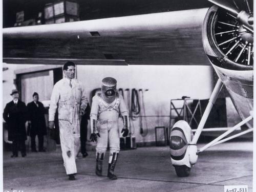 Wiley Post walks under his monoplane, wearing a pressure suit he planned to test while flying his aircraft. The pressure suit features a cylinder-shaped helmet and dark-colored boots.