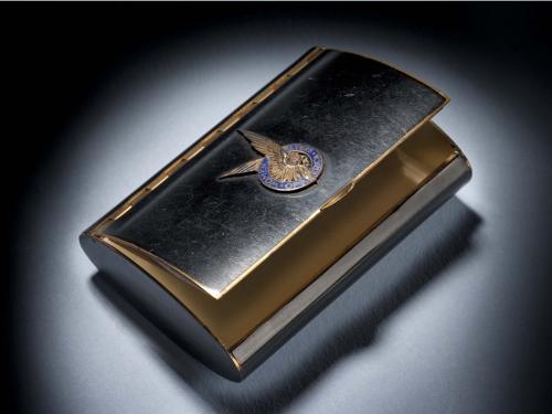 Black makeup case with open lid. On the center of the lid, there is a silver insignia of a pair of two wings inside a blue seal.