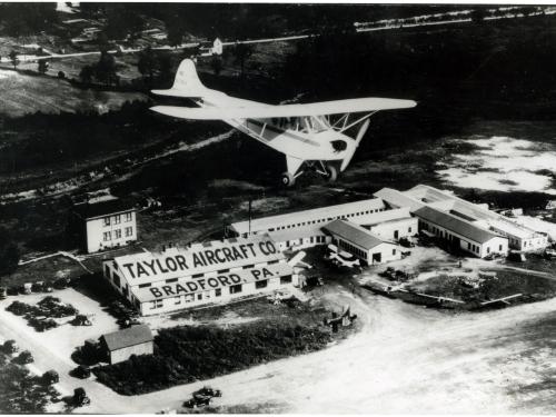 A monoplane flies over a set of buildings which are labeled as belonging to the "Taylor Aircraft Co." on the roof of a building on the left of the other buildings. Below the roof stating the company's name is the location of "Bradford, PA" on a lower roof.