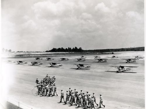 Multiple lines of Piper Cubs, monoplanes with one engine, are seen on a U.S. Navy training site. Groups of men are walking in formation in front of the planes.