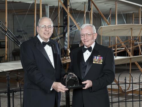 Two white men stand together, holding a trophy award at a Museum awards event.