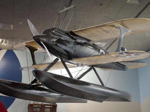 Diagonal and frontal view of black biplane with single engine and pontoon floats hanging in the Museum.