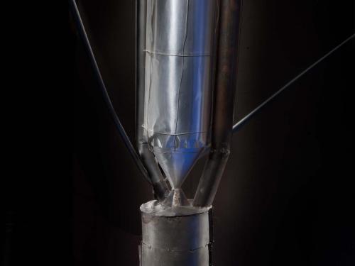 Close-up view of motor on a primitive rocket. A conical nose tops a metal cylinder-shaped motor attached to a cylinder-shaped fuel tank by a metal conical-shaped drain.