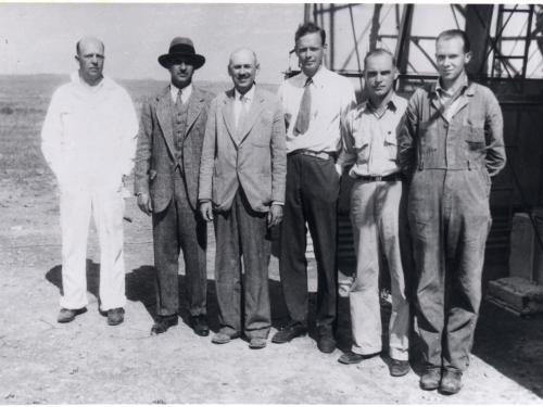 Six white men, including Harry Guggenheim, Robert Goddard, and Charles Lindbergh, stand in front of a tower in a desert.