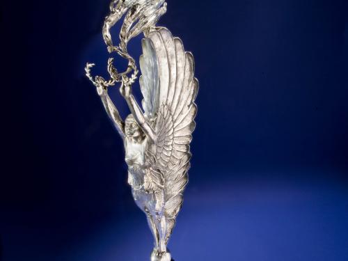 Silver-colored trophy with topper of a winged angel holding olive wreaths which are picked up by a plane.