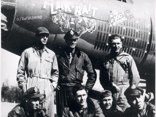 A group of seven members of a bomber combat plane used in World War II named "Flak-Bait" pose formally in front of the aircraft following a milestone mission.