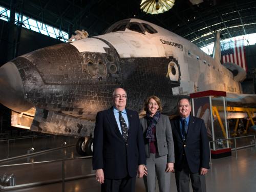 Three people, two white men and one white woman, stand formally in front of the Space Shuttle Discovery, a white spacecraft.