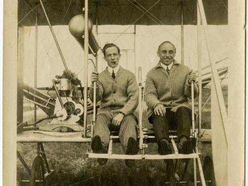 Harry Bingham Brown, a white male pilot, sits in his Wright biplane on the left of another male. Brown is smoking a cigar.