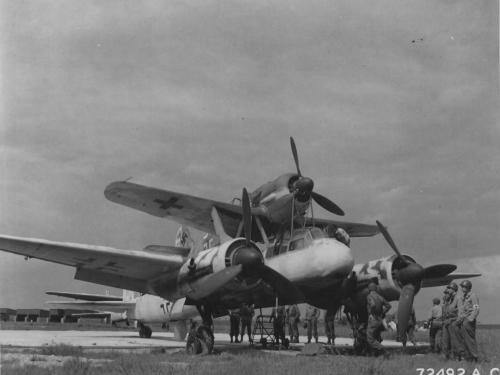 Men stand around a aircraft with two engines which is holding a smaller monoplane with one engine on top of the fuselage.