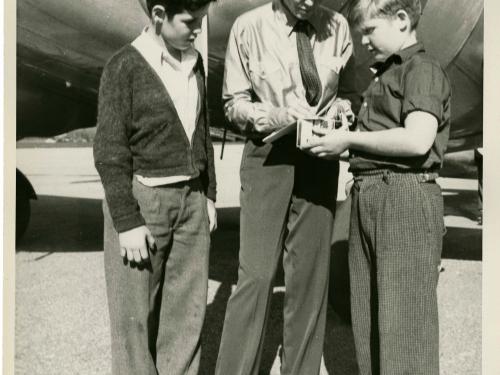 Amelia Earhart, a white woman, stands in between two white people in front of an aircraft. She is signing an autograph for the person on her left (our right).