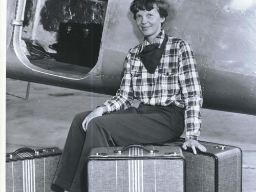 Amelia Earhart, a white female pilot, poses in front of an aircraft with three pieces of luggage that she designed.