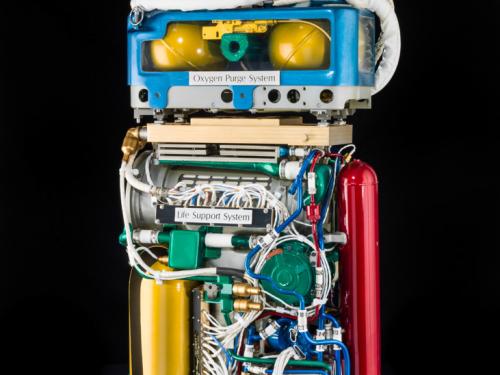 Cutaway view of life support system, a large set of wires and tubes attached to multiple cylinders, and oxygen purge system, which features multiple smaller cylinders around a green tube.