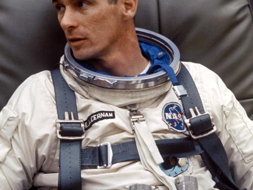 Astronaut Eugene Cernan, a white male, sits while wearing his astronaut suit without the helmet. Multiple life support tubes are seen attached to the suit.