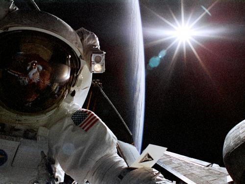 Astronaut Joseph R. Tanner, a white male astronaut, poses during a spacewalk. A silhouette of the Earth can be seen behind him.