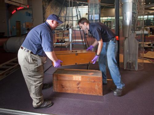 Two Museum staff members work to move a set of wooden boxes out of an exhibit.