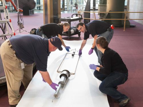 Four Museum staff members work to prepare a primitive rocket for travel away from a museum exhibit.