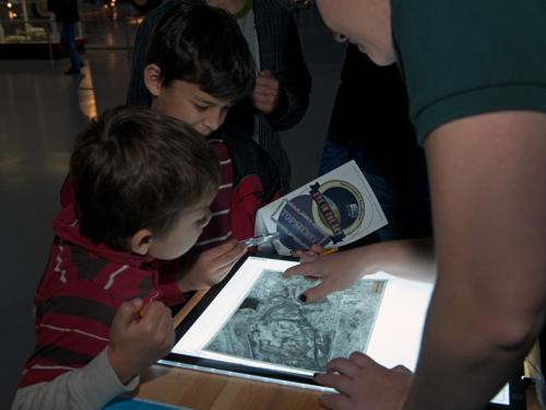 One staff member of the Museum and two younger-aged Museum visitors work together to solve a game. They are looking at an object on a white screen.