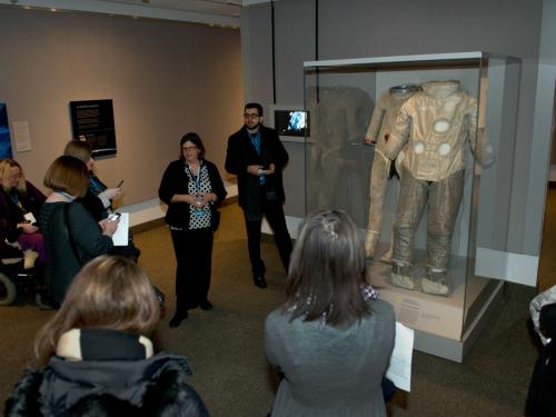 A white female museum conservator speaks to visitors about two tan brown-colored spacesuits on display inside an exhibit about spaceflight.