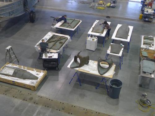 Multiple pieces of a gray, metal aircraft are placed separately on eight tables. Three Museum conservators are performing conservation treatments on separate pieces of the aircraft.